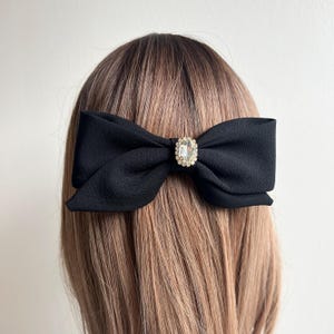 May include: A black fabric bow hair clip with a sparkling, oval-shaped rhinestone center. The bow is attached to a metal clip, designed to be worn in the hair. The hair accessory is shown on a person with light brown hair.