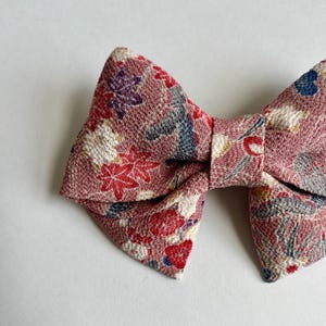 May include: A bow-shaped hair accessory made of textured fabric in a dusty rose pink hue. The material features a detailed pattern of small red, white, purple, and blue floral and leaf motifs scattered across the entire surface of the bow.
