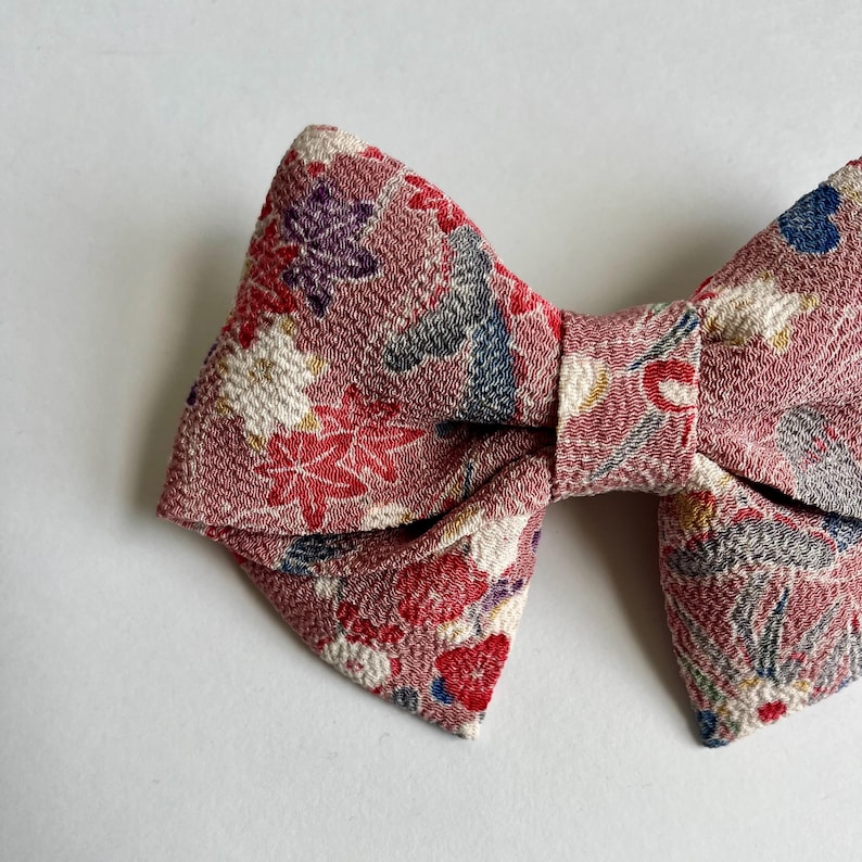 May include: A bow-shaped hair accessory made of textured fabric in a dusty rose pink hue. The material features a detailed pattern of small red, white, purple, and blue floral and leaf motifs scattered across the entire surface of the bow.