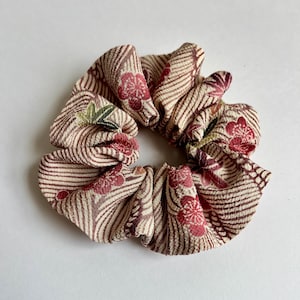 May include: A fabric hair scrunchie featuring a beige background with a pattern of dark brown diagonal stripes and red floral motifs. The accessory has a gathered, ruffled texture throughout, creating a circular shape with a hollow center.