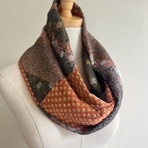 Puede incluir: Una bufanda infinita con un diseño de patchwork de varias telas en tonos naranja quemado, verde oliva, marrón y negro. Los patrones incluyen pequeños cuadrados geométricos, motivos florales intrincados y detalles de líneas en estrella.