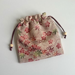 May include: A drawstring pouch featuring a beige fabric base with a wavy line pattern, decorated with red plum blossoms and green and red maple leaves. The bag includes dark maroon cord strings finished with small, light-colored wooden beads.