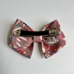May include: A hair clip featuring a bow made of textured fabric in a dusty rose color. The fabric displays a pattern of small red, blue, and white floral and leaf motifs. A silver-toned metal barrette mechanism is attached to the back of the bow.