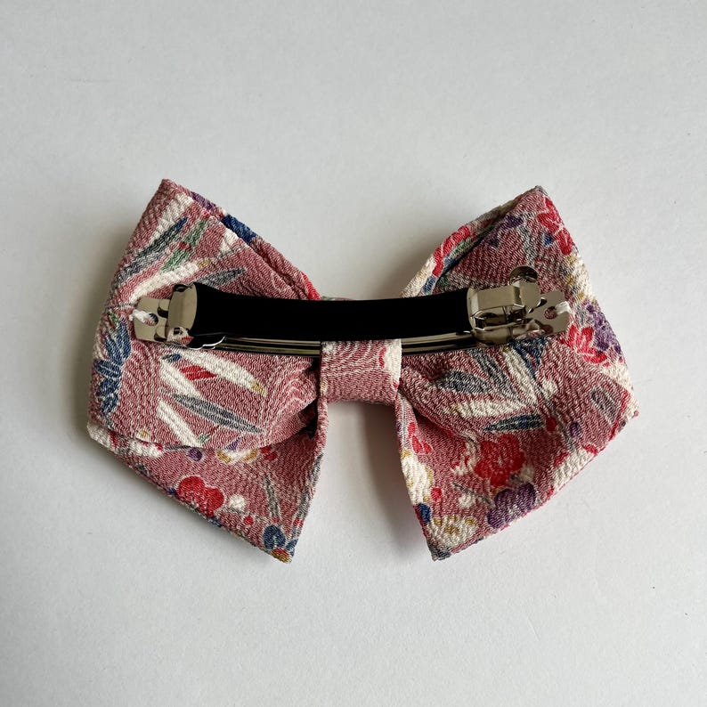 May include: A hair clip featuring a bow made of textured fabric in a dusty rose color. The fabric displays a pattern of small red, blue, and white floral and leaf motifs. A silver-toned metal barrette mechanism is attached to the back of the bow.