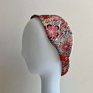 May include: A wide fabric headband featuring a vibrant red base with a detailed floral pattern in white, gold, teal, and black. The textured material displays stylized blossoms and geometric accents, with a gathered elastic section at the base.