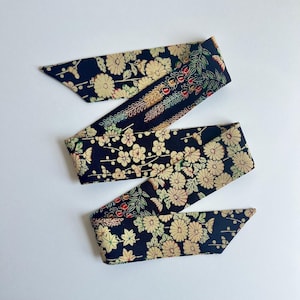 May include: A long, narrow fabric ribbon featuring a dark navy blue background with a repeating pattern of cream-colored chrysanthemum flowers, green leaves, and small red berries. The ribbon has pointed ends and a smooth, flat texture throughout.