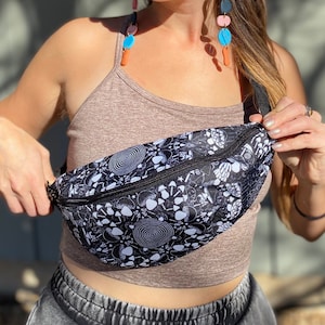 May include: A black fanny pack with a white psychedelic print featuring mushrooms, skulls, and other symbols. The fanny pack is worn by a person with long brown hair and colorful earrings.