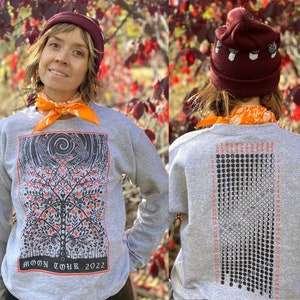 K&ouml;nnte beinhalten: Ein graues Sweatshirt mit einer schwarz-wei&szlig;en Illustration eines Baumes mit V&ouml;geln und einem Mond auf der Vorderseite. Die R&uuml;ckseite des Sweatshirts zeigt eine schwarz-wei&szlig;e Illustration eines Mondkalenders mit dem Text "MOON TOUR 2022".
