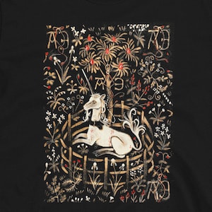Op de afbeelding: Zwart T-shirt met een wit eenhoorn in een cirkelvormig frame, omringd door een bloemenpatroon. Het eenhoorn ligt met zijn hoofd naar de zijkant gedraaid. Het bloemenpatroon is in tinten bruin, rood en geel.
