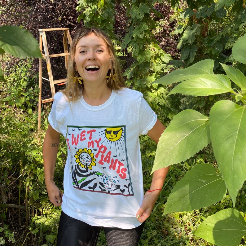 May include: A white t-shirt with a graphic of a cartoon sunflower wearing sunglasses and watering plants. The text on the shirt reads "I Wet My Plants".