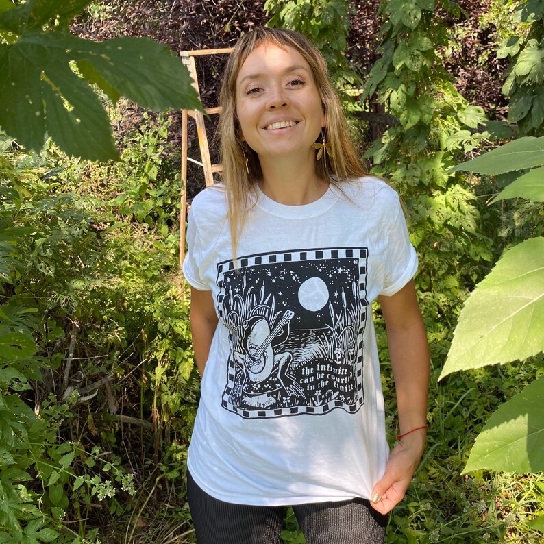 May include: White t-shirt with a black and white illustration of a frog playing a guitar under a full moon. The text "The infinite can be found in the finite" is printed below the frog.
