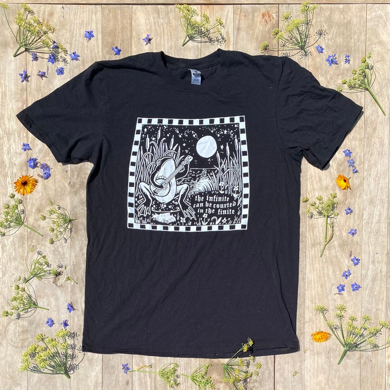 May include: Black t-shirt featuring a white graphic of a frog playing a banjo in a moonlit swamp. The text "the infinite can be counted in the finite" is below the frog.
