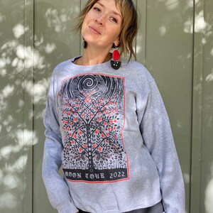 K&ouml;nnte beinhalten: Ein graues Sweatshirt mit einem schwarzen und roten Grafikdruck eines Baumes mit V&ouml;geln und dem Text "MOON TOUR 2022".