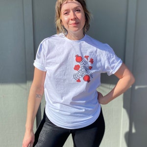 May include: A white t-shirt with a red pomegranate design. The design features a branch with leaves and multiple pomegranates.
