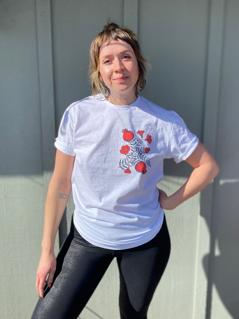 May include: A white t-shirt with a red pomegranate design. The design features a branch with leaves and multiple pomegranates.