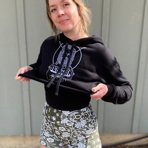 May include: A black cropped hoodie with a white graphic of a face and the word "Jupiter" on the front. The model is wearing the hoodie with a pair of green and white floral print shorts.