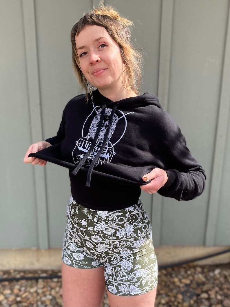 May include: A black cropped hoodie with a white graphic of a face and the word "Jupiter" on the front. The model is wearing the hoodie with a pair of green and white floral print shorts.