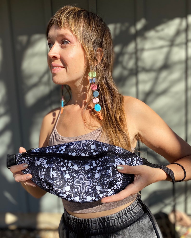 May include: A black and white fanny pack with a psychedelic mushroom and spiral pattern. The fanny pack has a black zipper and a black adjustable strap.