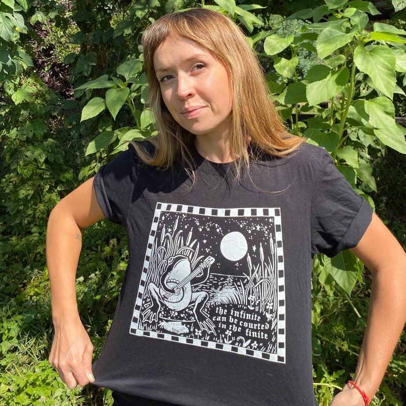 May include: Black t-shirt featuring a white graphic of a frog playing a guitar under a full moon. The text "The infinite can be courted in the finite" is printed below the frog.