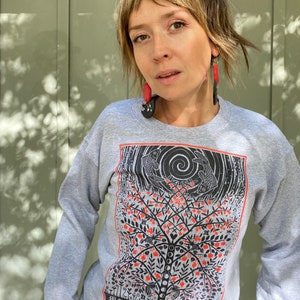 K&ouml;nnte beinhalten: Ein graues Sweatshirt mit einem schwarzen und orangenen Druck eines Baumes mit roten &Auml;pfeln und zwei Fischen. Der Text "MOON TOUR 2022" ist unten auf dem Design gedruckt.