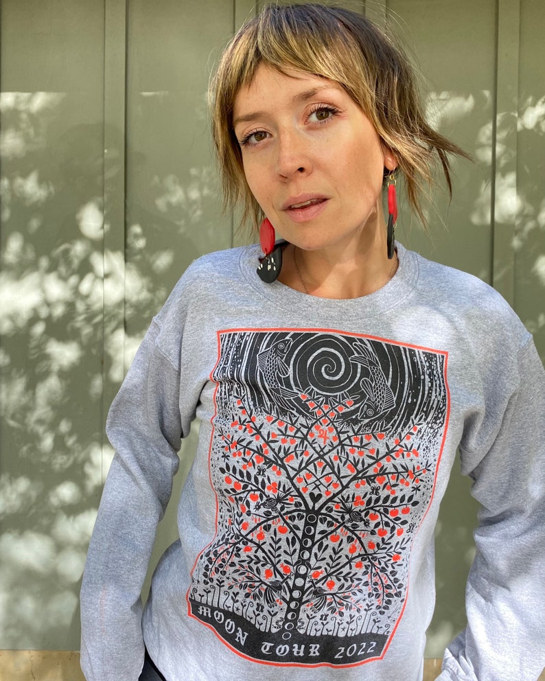 K&ouml;nnte beinhalten: Ein graues Sweatshirt mit einem schwarzen und orangenen Druck eines Baumes mit roten &Auml;pfeln und zwei Fischen. Der Text "MOON TOUR 2022" ist unten auf dem Design gedruckt.
