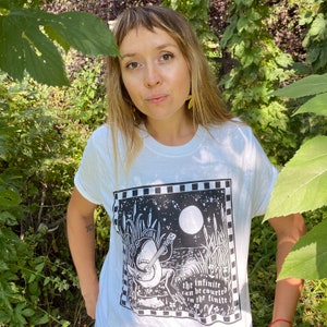 May include: White t-shirt with a black and white illustration of a frog playing a guitar in a moonlit swamp. The text "the infinite can be counted in the finite" is printed below the frog.
