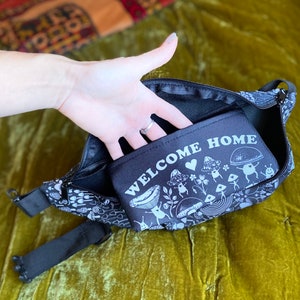 May include: Black fanny pack with a white mushroom design and the text "Welcome Home". The fanny pack has a zipper closure and a small pocket on the front.