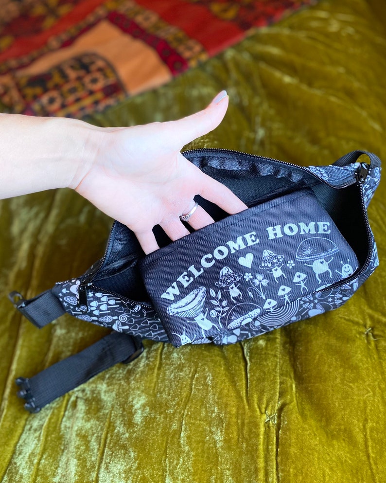 May include: Black fanny pack with a white mushroom design and the text "Welcome Home". The fanny pack has a zipper closure and a small pocket on the front.
