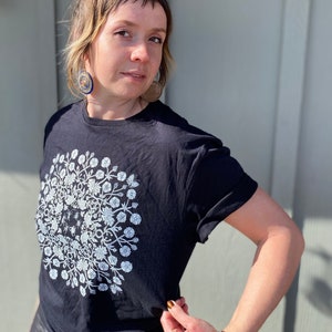 May include: A black t-shirt with a white floral design. The design is a circle with a floral pattern and a small bee in the center.