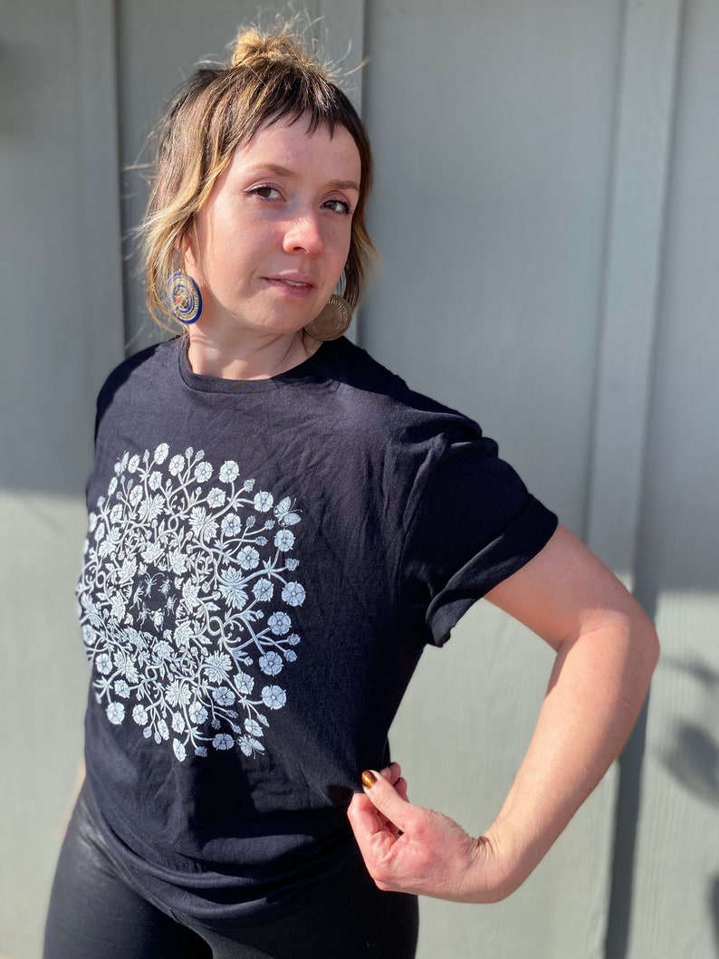 May include: A black t-shirt with a white floral design. The design is a circle with a floral pattern and a small bee in the center.