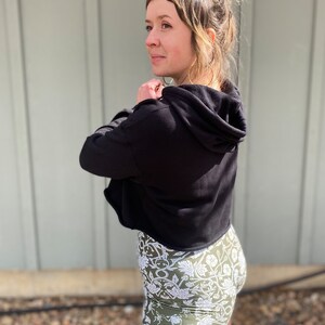 May include: A woman wearing a black cropped hoodie with a hood and a pair of green and white floral print shorts.