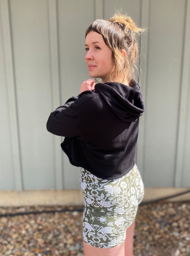 May include: A woman wearing a black cropped hoodie with a hood and a pair of green and white floral print shorts.