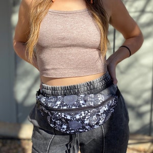 May include: A black and white fanny pack with a psychedelic mushroom and spiral design. The fanny pack is worn over a brown crop top and black sweatpants.