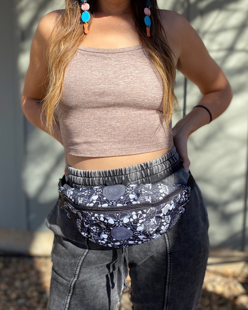 May include: A black and white fanny pack with a psychedelic mushroom and spiral design. The fanny pack is worn over a brown crop top and black sweatpants.