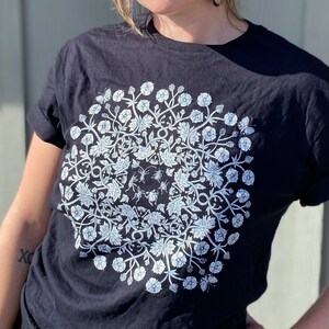May include: Black t-shirt with a white floral design featuring a circle of flowers, leaves, and insects.