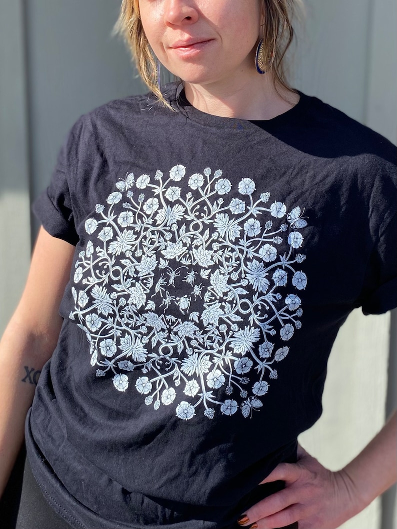 May include: Black t-shirt with a white floral design featuring a circle of flowers, leaves, and insects.