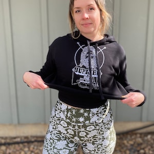 May include: A black cropped hoodie with a white graphic print of a person with a beard and the text "Jupiter is my Homeboy". The person is wearing a black hoodie with a white graphic print of a person with a beard and the text "Jupiter is my Homeboy". The person is wearing a pair of green and white floral print shorts.