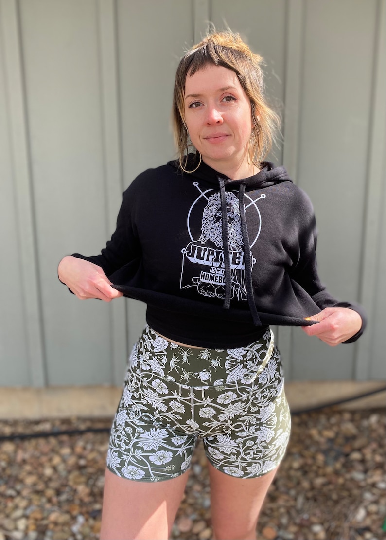 May include: A black cropped hoodie with a white graphic print of a person with a beard and the text "Jupiter is my Homeboy". The person is wearing a black hoodie with a white graphic print of a person with a beard and the text "Jupiter is my Homeboy". The person is wearing a pair of green and white floral print shorts.