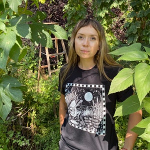 May include: A person wearing a black t-shirt with a white graphic print of a creature playing a banjo. The graphic includes the text "The infinite can be counted in the infinite".