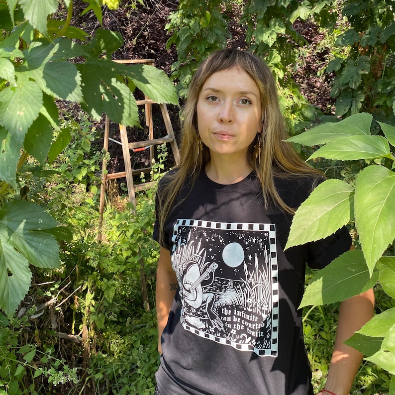 May include: A person wearing a black t-shirt with a white graphic print of a creature playing a banjo. The graphic includes the text "The infinite can be counted in the infinite".