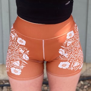 May include: Brown athletic shorts with a white floral design. The shorts are high-waisted and have a V-shaped waistband. The floral pattern is symmetrical, with white flowers and stems on each side of the shorts.