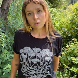 K&ouml;nnte beinhalten: Ein schwarzes T-Shirt mit einem wei&szlig;en Aufdruck einer Gruppe von Schafgarbenbl&uuml;ten. Der Aufdruck ist detailliert und beinhaltet die Wurzeln der Pflanze. Das Shirt wird von einer Person mit langen braunen Haaren getragen.