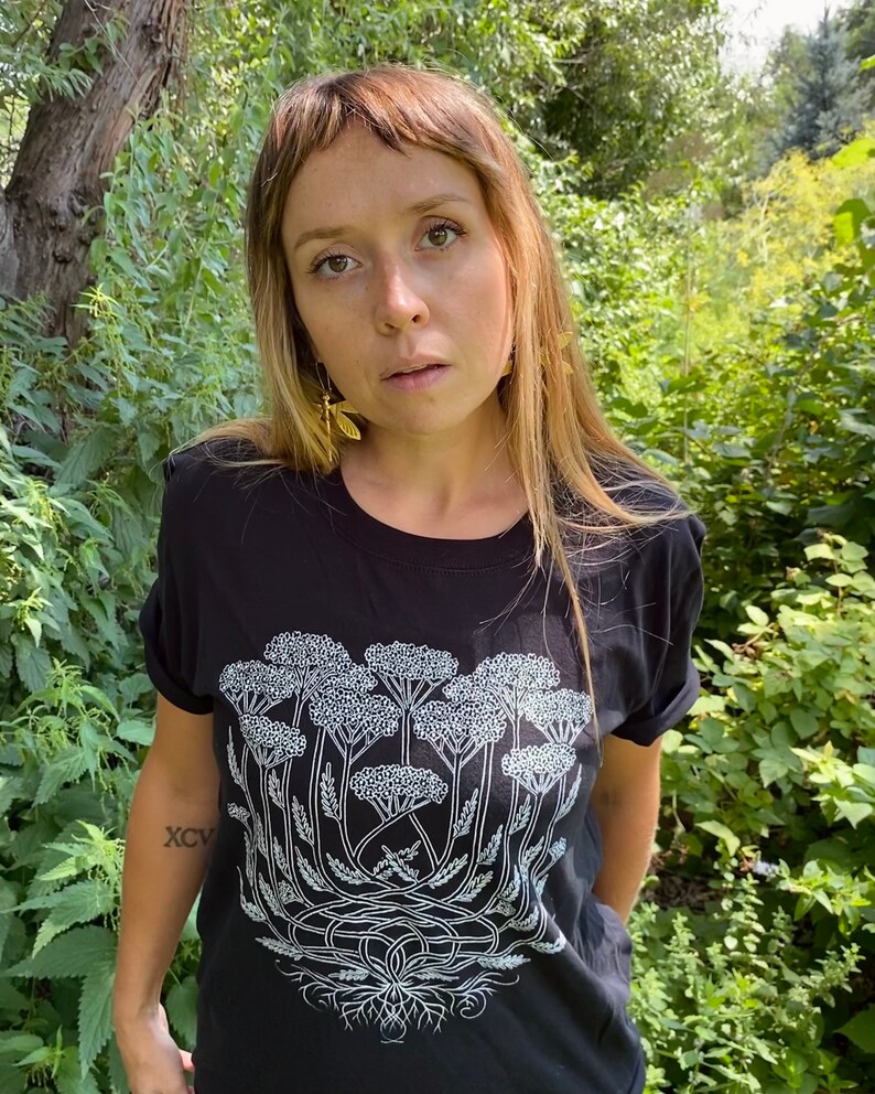 K&ouml;nnte beinhalten: Ein schwarzes T-Shirt mit einem wei&szlig;en Aufdruck einer Gruppe von Schafgarbenbl&uuml;ten. Der Aufdruck ist detailliert und beinhaltet die Wurzeln der Pflanze. Das Shirt wird von einer Person mit langen braunen Haaren getragen.