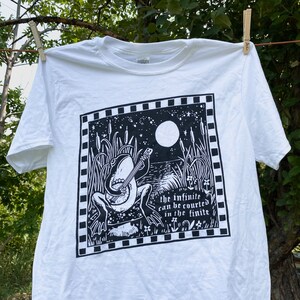 May include: White t-shirt with a black and white illustration of a frog playing a banjo in a swamp setting under a full moon. The text "the infinite can be courted in the finite" is printed below the frog.