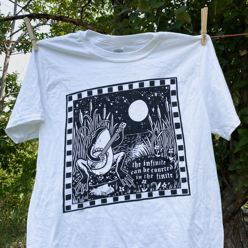 May include: White t-shirt with a black and white illustration of a frog playing a banjo in a swamp setting under a full moon. The text "the infinite can be courted in the finite" is printed below the frog.