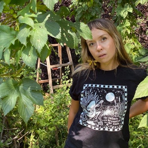 May include: A person wearing a black t-shirt with a white graphic of a frog playing a guitar. The graphic includes the text "The infinite can be counted in the finite".