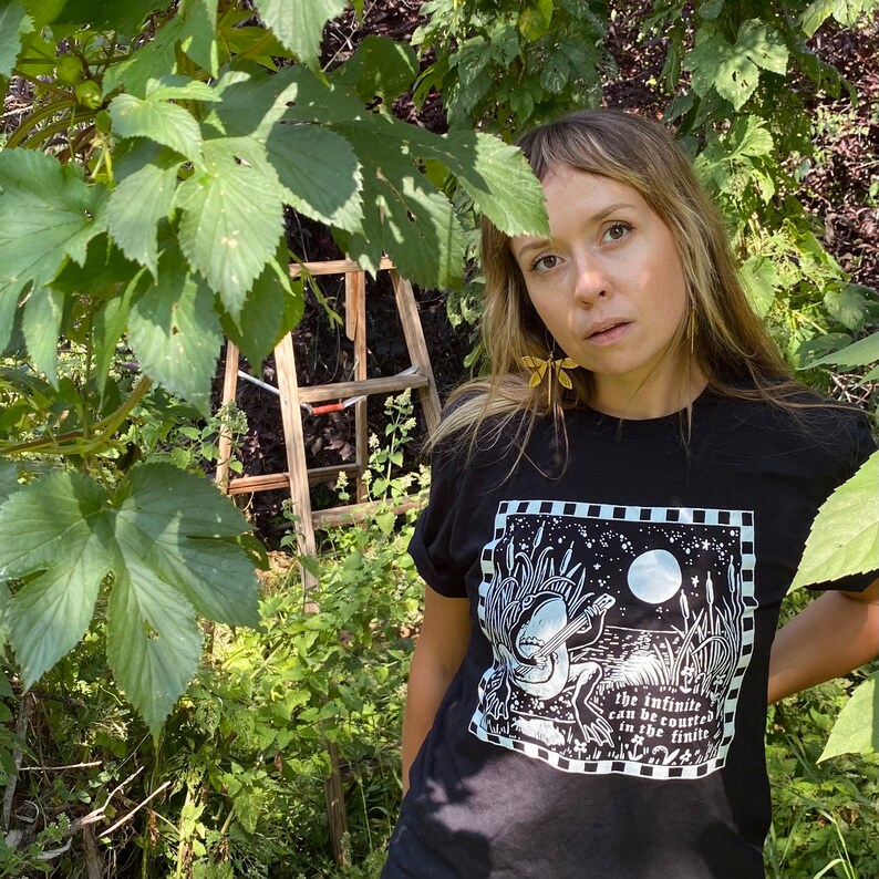May include: A person wearing a black t-shirt with a white graphic of a frog playing a guitar. The graphic includes the text "The infinite can be counted in the finite".