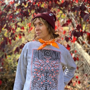 K&ouml;nnte beinhalten: Ein graues Sweatshirt mit einer Schwarz-Wei&szlig;-Illustration eines Baumes mit roten &Auml;pfeln. Der Baum ist von einem schwarzen Rahmen mit dem Text "MOON TOUR 2022" umgeben. Die Person, die das Sweatshirt tr&auml;gt, tr&auml;gt auch eine kastanienbraune M&uuml;tze und ein orangefarbenes Bandana.