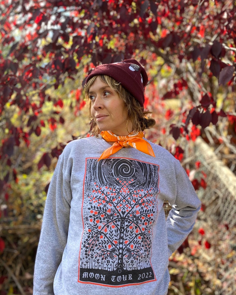 K&ouml;nnte beinhalten: Ein graues Sweatshirt mit einer Schwarz-Wei&szlig;-Illustration eines Baumes mit roten &Auml;pfeln. Der Baum ist von einem schwarzen Rahmen mit dem Text "MOON TOUR 2022" umgeben. Die Person, die das Sweatshirt tr&auml;gt, tr&auml;gt auch eine kastanienbraune M&uuml;tze und ein orangefarbenes Bandana.