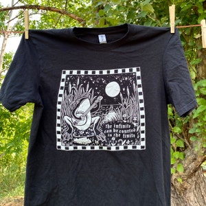 May include: Black t-shirt featuring a white graphic print of a frog playing a banjo under a full moon. The text "the infinite can be courted in the finite" is printed below the frog.